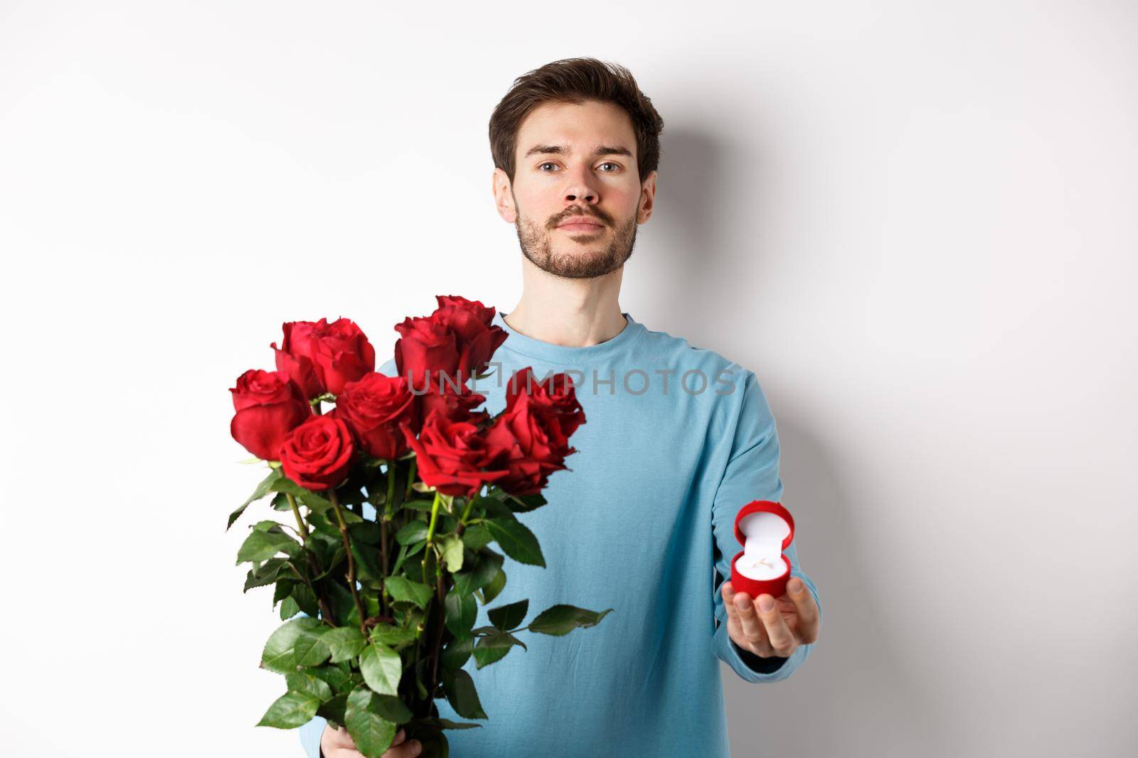 Valentines and relationship. Romantic man boyfriend holding red roses and showing engagement ring, making a proposal on lovers day, standing over white background.