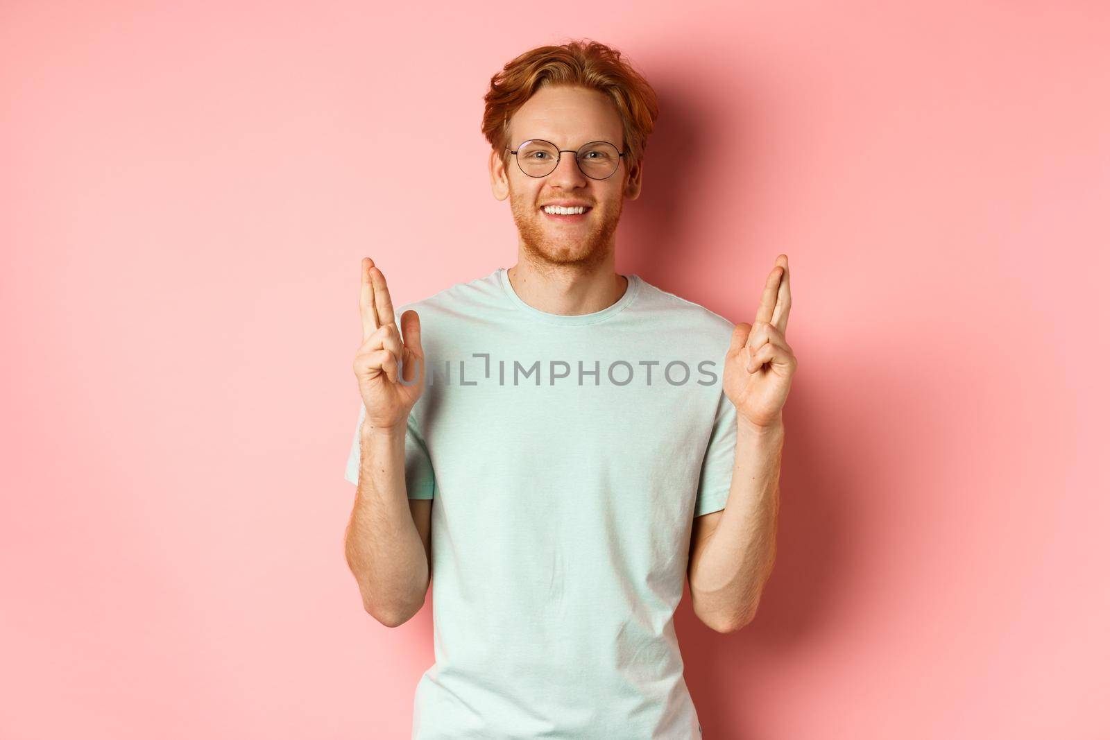 Attractive guy having faith in dreams, smiling hopeful and making wish with fingers crossed, feeling lucky, standing over pink background.