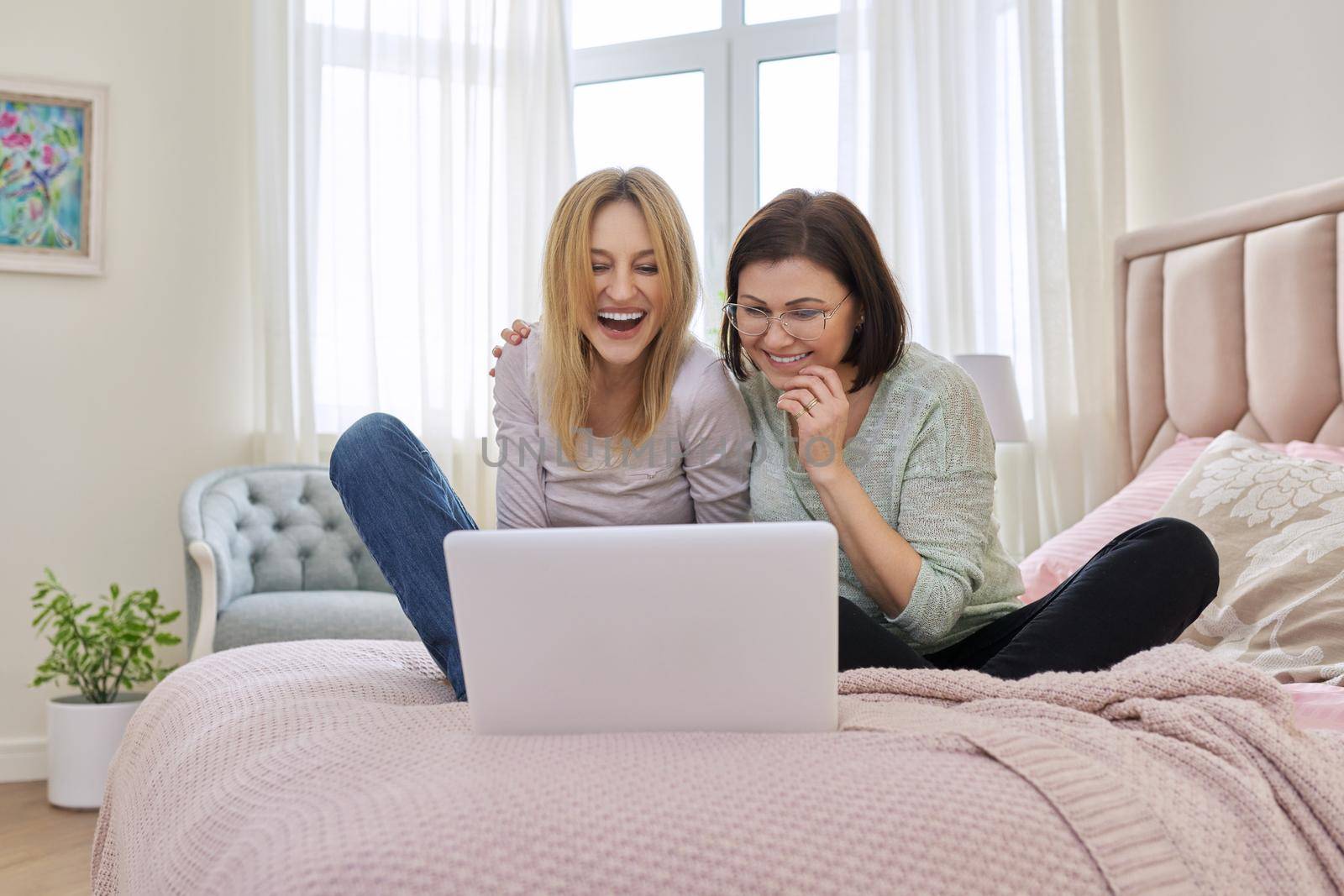 Two happy middle aged women having rest sitting together at home on bed, looking in laptop screen. Same sex female couple, relationship, lifestyle, people concept
