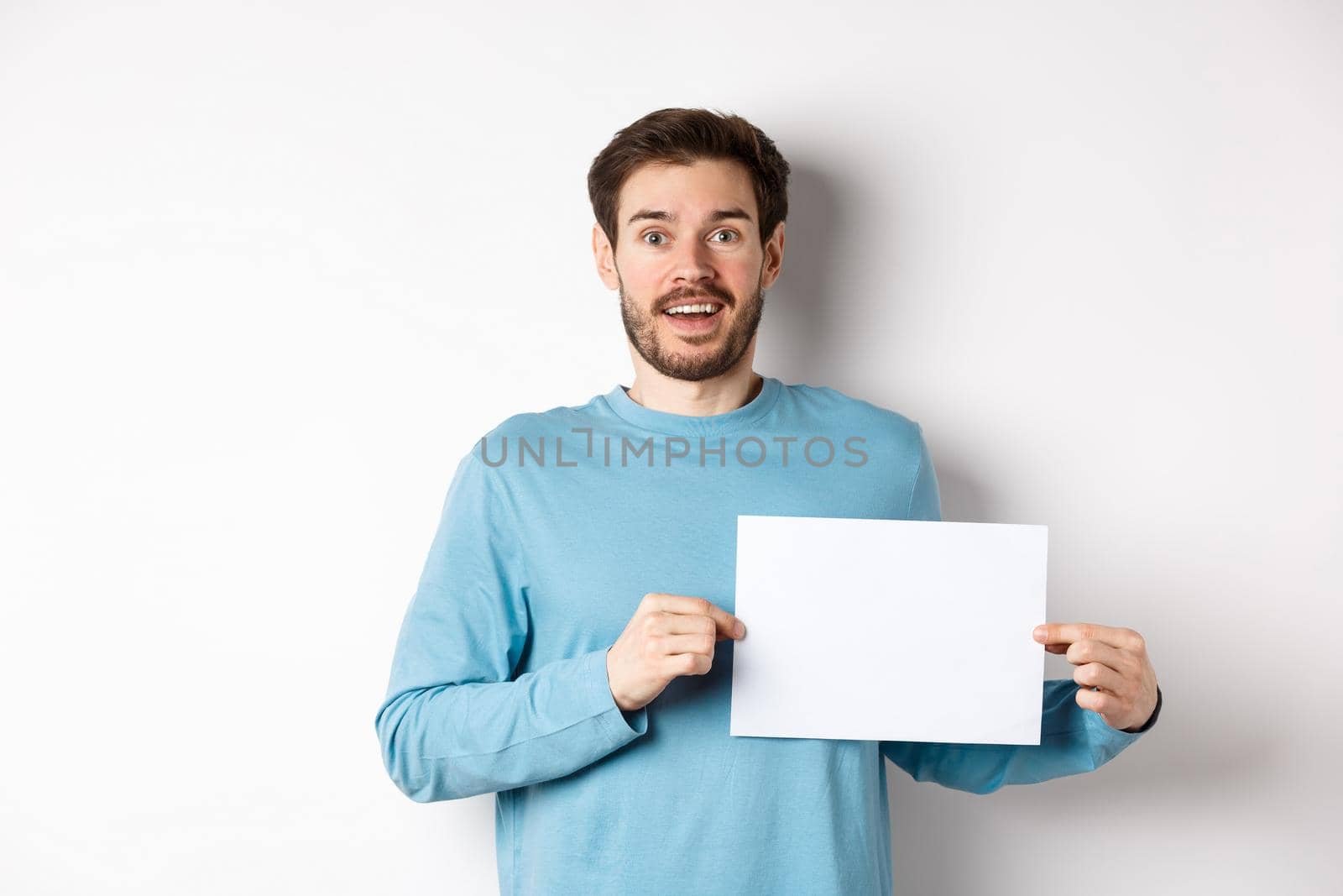 Romantic man looking with hopeful face at camera, showing sign card, blank piece of paper for banner or logo, standing over white background by Benzoix