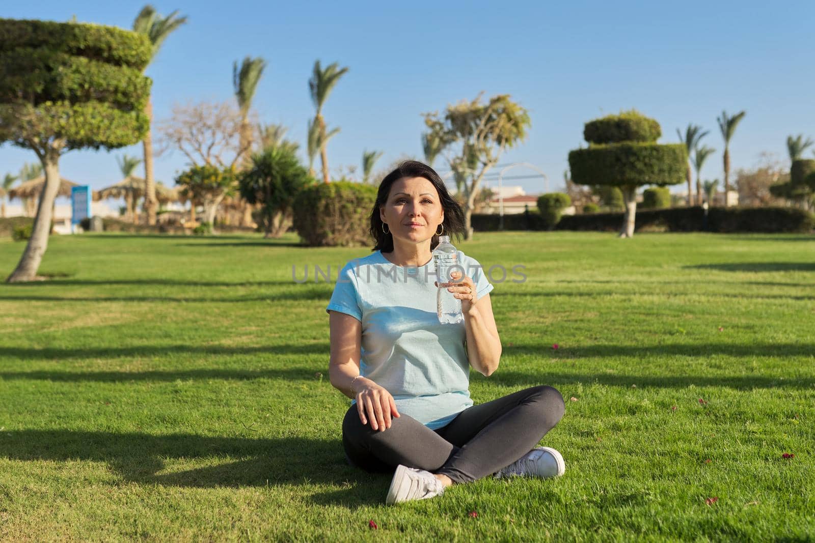 Mature woman doing morning exercises on green grass in park, with bottle of water by VH-studio