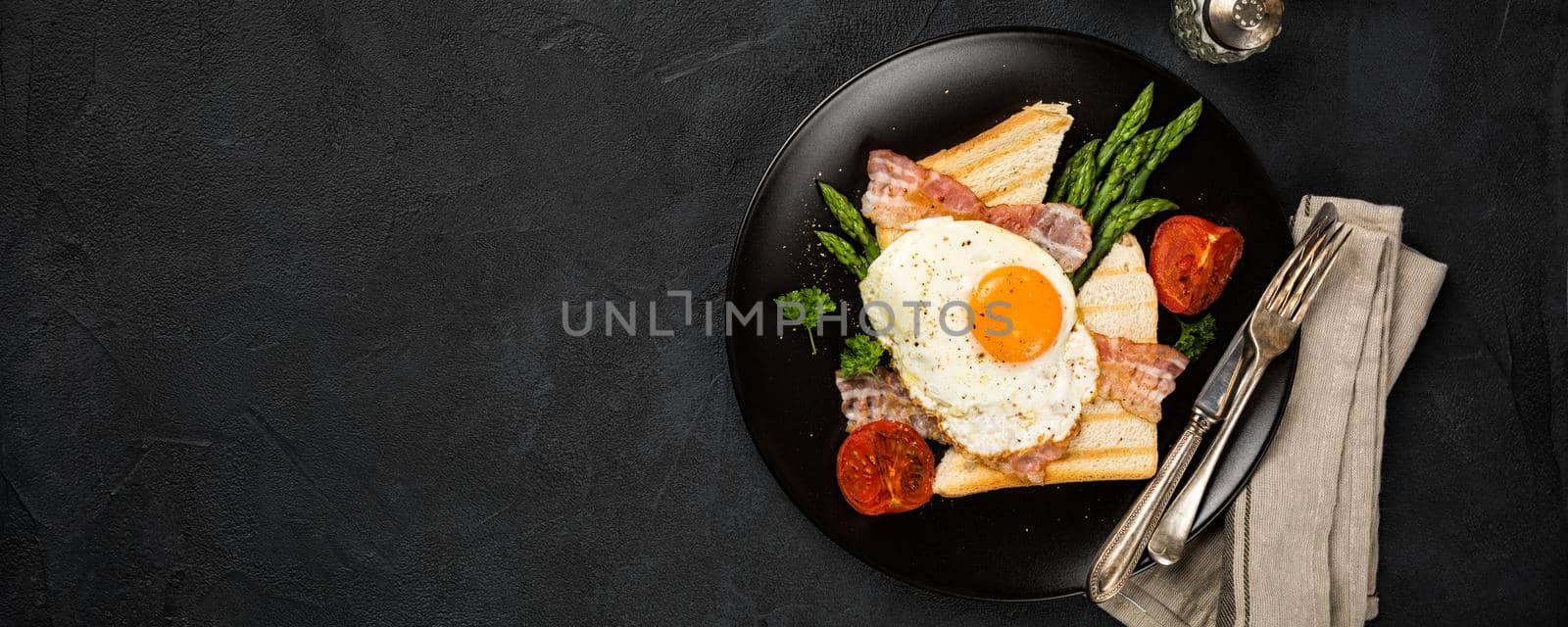 Breakfast or lunch with Fried egg, bread toast, green asparagus, tomatoes and bacon on black plate. Top view. Copy space. Banner.