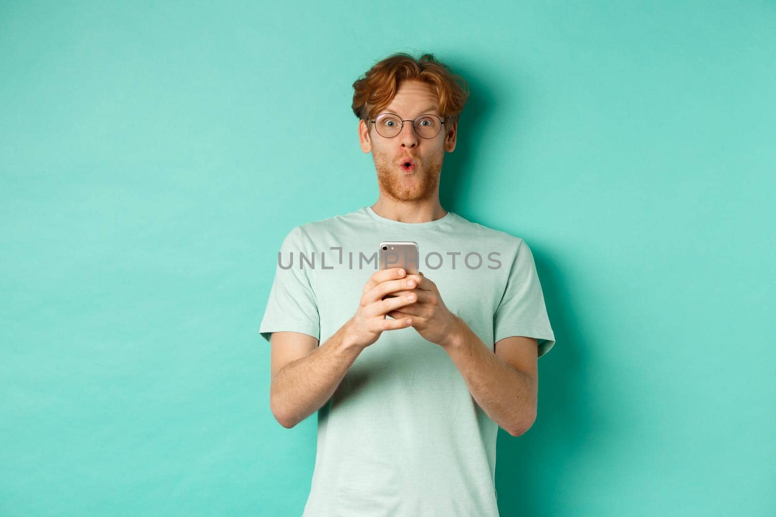 Impressed young man with red hair and glasses, holding mobile phone, saying wow and stare amazed at camera, standing over mint background.