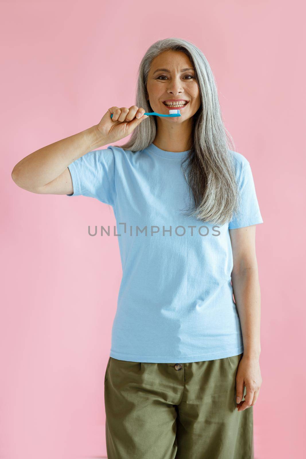 Positive long haired mature Asian model holds toothbrush on pink background by Yaroslav_astakhov