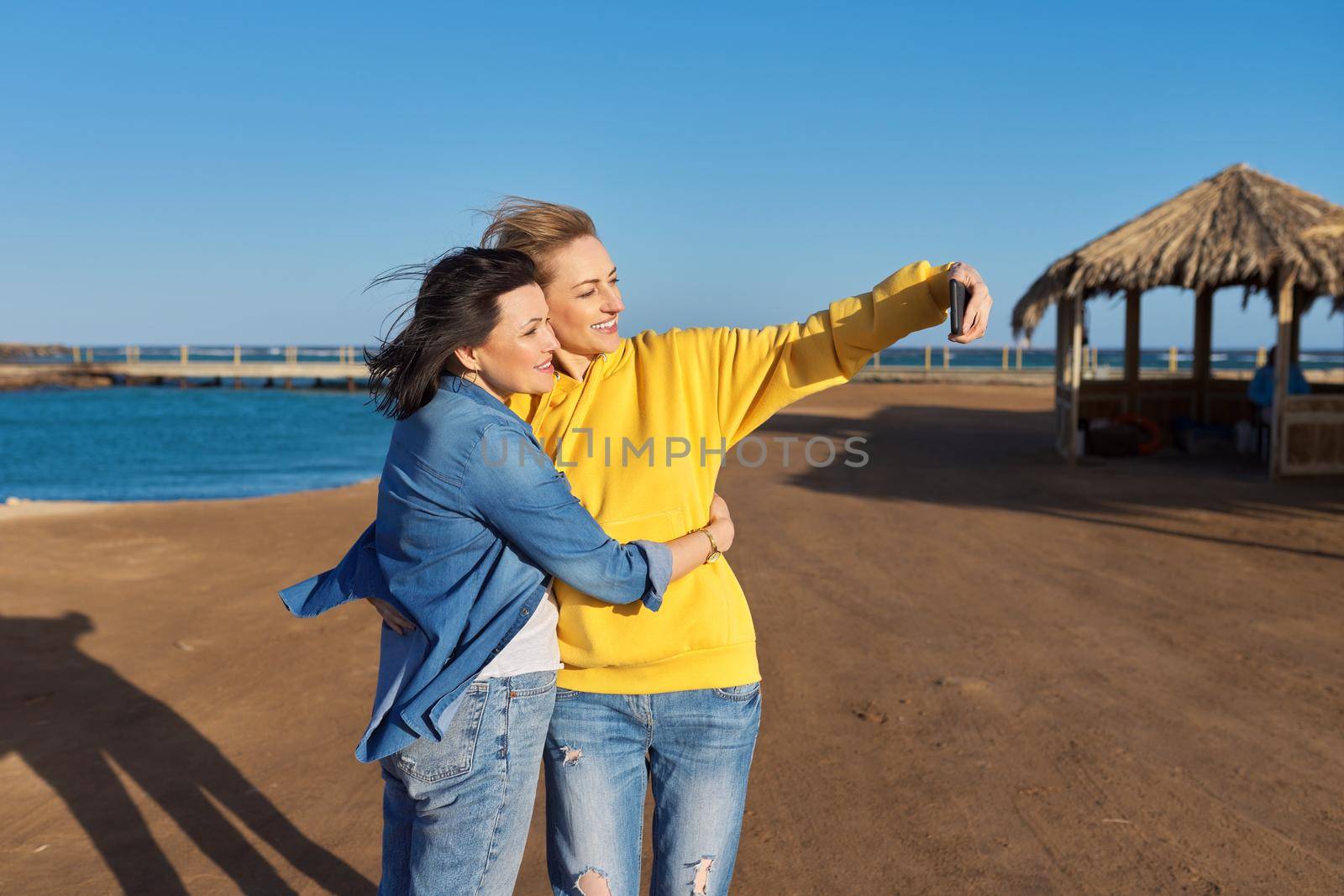 Two mature happy women taking selfie photo together using smartphone. Sea beach, resort, nature background. Friendship, family, same-sex couple, vacation, weekend, relaxation at sea concept