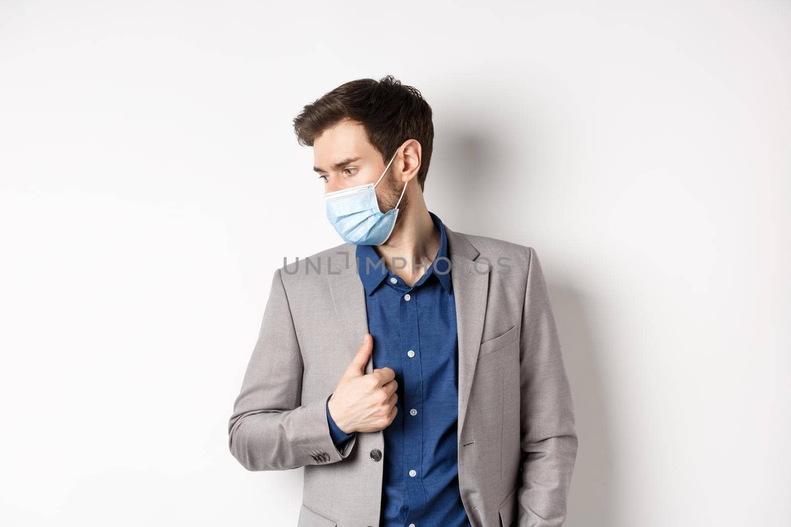 Covid, pandemic and business concept. Handsome male entrepreneur wearing stylish suit and medical mask, looking aside, white background.