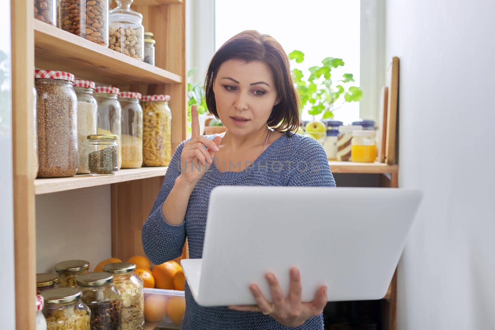 Woman in kitchen pantry with stored products, holding laptop by VH-studio