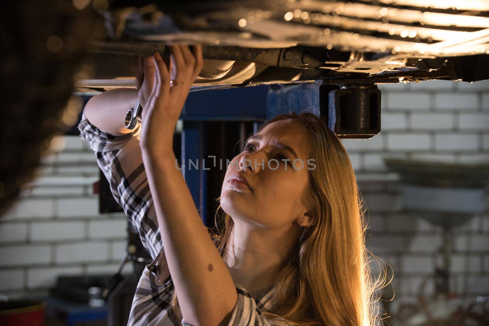 Car repair service. Young sexy mechanic woman standing under the car holding a wrench and looking up. Mid shot