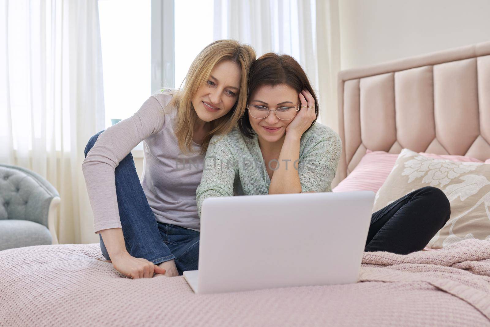 Two happy middle aged women having rest sitting together at home on bed, looking in laptop screen. Same sex female couple, relationship, lifestyle, people concept