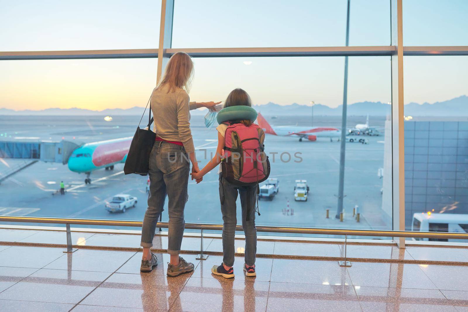 Airport passengers, standing with their backs family mother and daughter child looking at planes in panoramic window, awaiting boarding, copy space