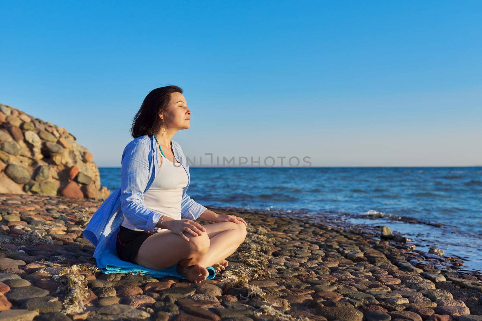 Beautiful middle aged woman sitting in lotus position and meditating on the seashore at sunset. Relaxation, relaxation, enjoying nature by the sea