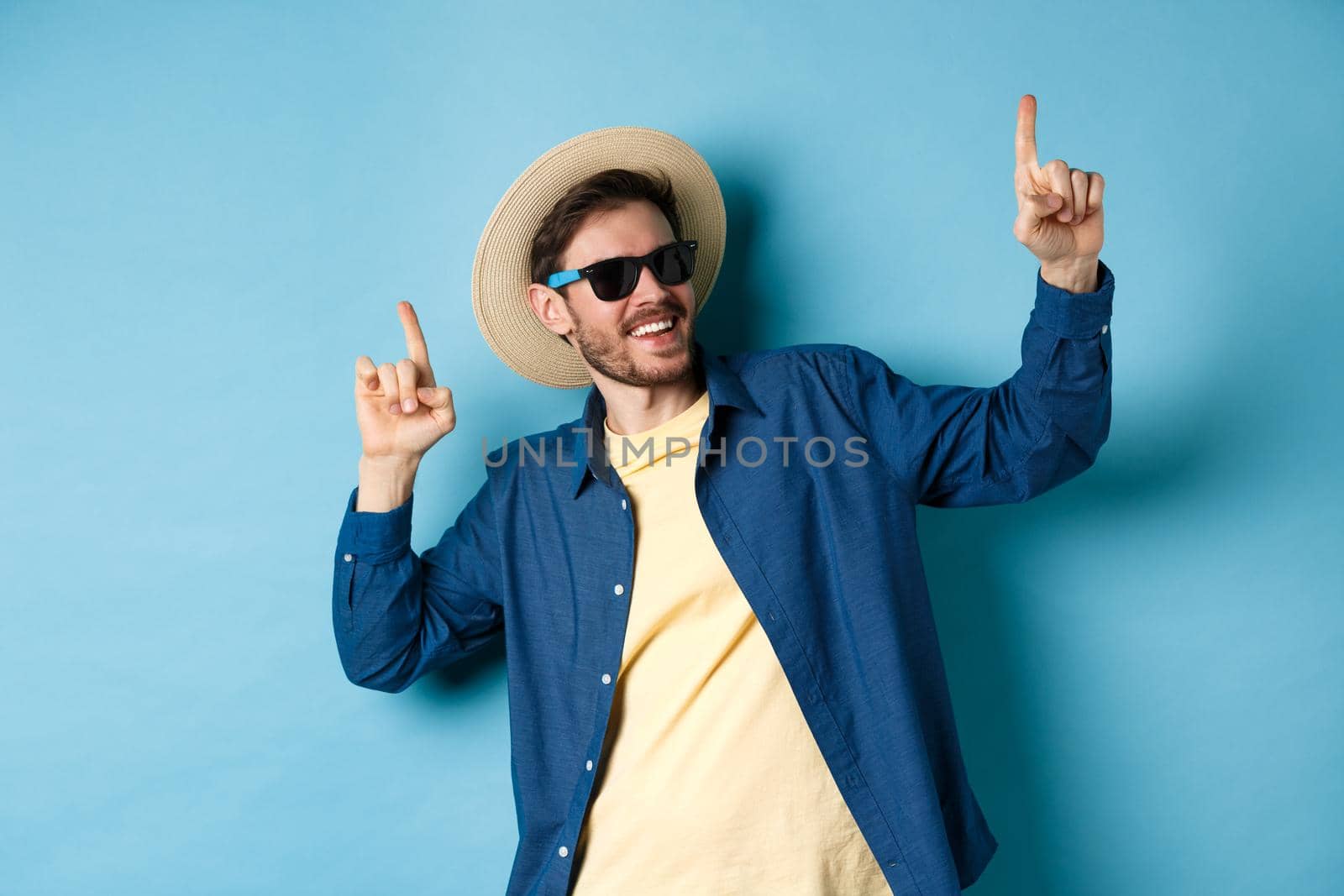 Funny tourist dancing on vacation, pointing fingers up, wearing summer hat and sunglasses, blue background.