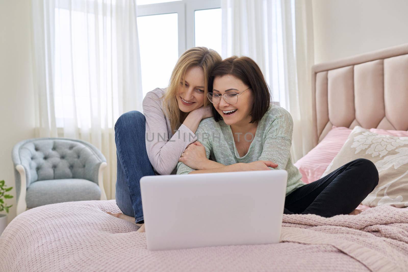 Two happy middle aged women having rest sitting together at home on bed, looking in laptop screen. Same sex female couple, relationship, lifestyle, people concept