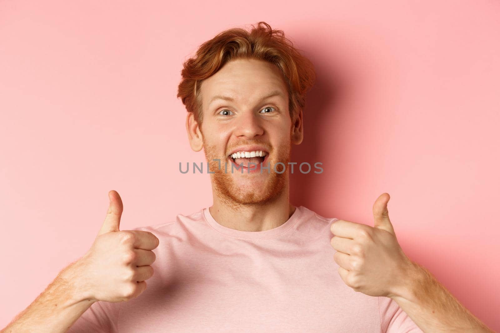 Close up of cheerful man with red hair and beard, showing thumbs up and smiling, saying yes, approve and praise something cool, standing over pink background.