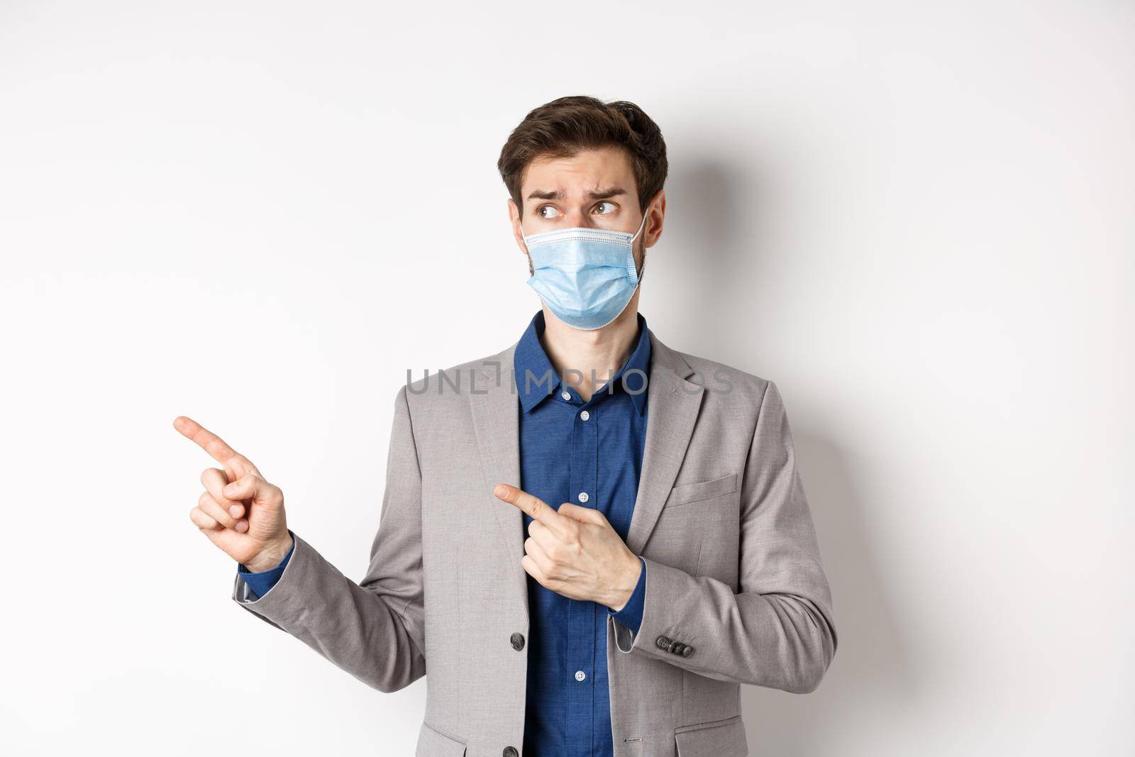 Covid-19, pandemic and business concept. Hesitant businessman in medical mask and suit looking, pointing left with doubtful face, standing on white background.