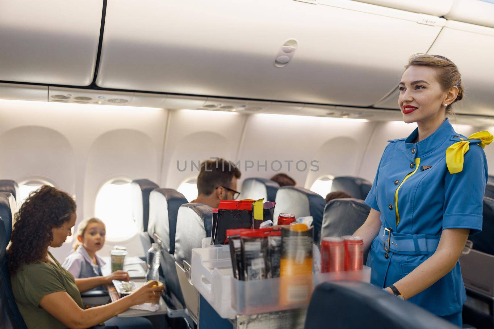 Female flight attendant serving food to passengers on aircraft. Hostess walking with trolley on aisle by Yaroslav_astakhov