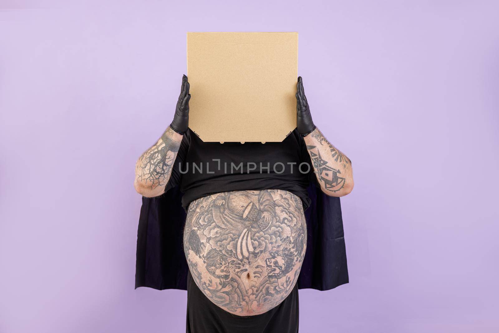 Plus size man wearing hero suit with tattoos on large bare tummy holds blank cardboard boxes of pizza posing on purple background in studio