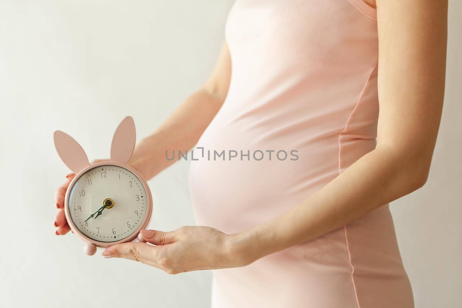 Closeup of a pregnant woman in pink dress holding bunny shaped alarm clock near her belly-maternity concept on white background