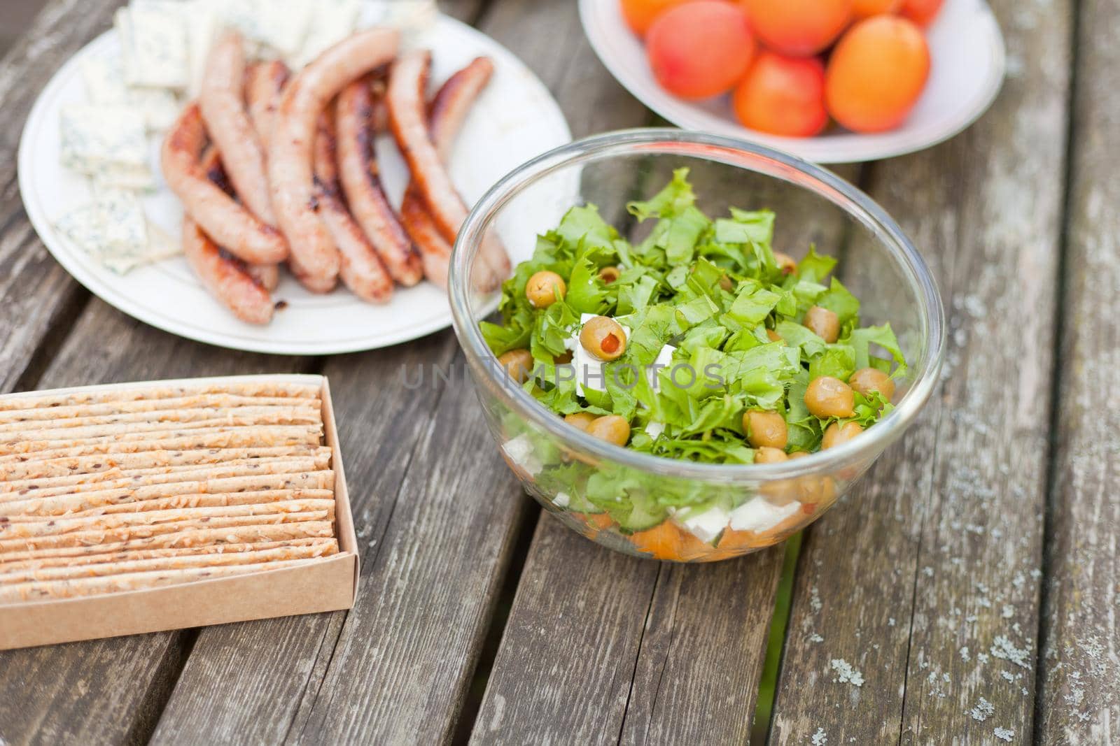 Fresh healthy salad with olives and feta chees on wooden table. Baked crisps with sausages at the picnic time. Tasty fresh food