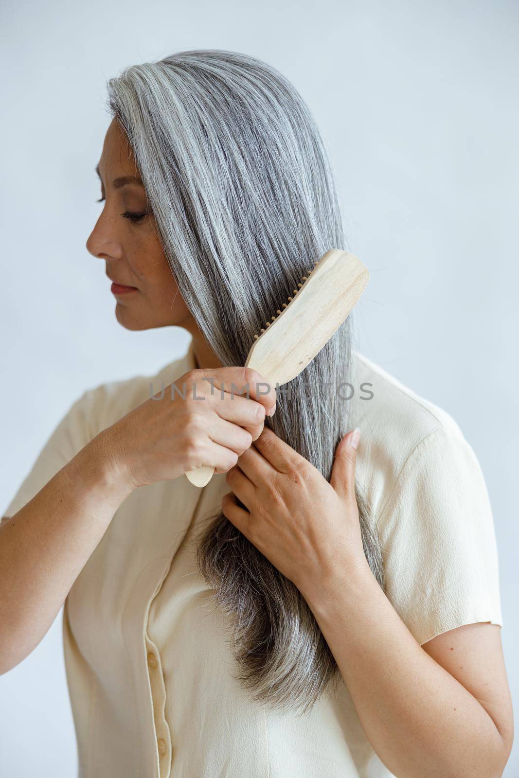 Languid middle aged Asian woman brushes natural hoary hair posing on light grey background in studio. Mature beauty lifestyle