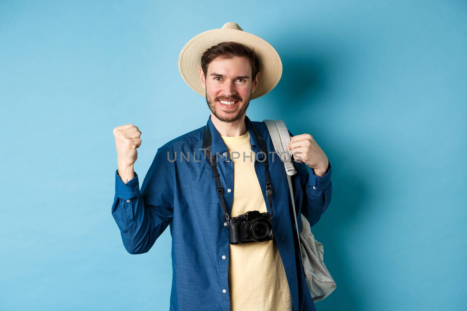 Happy tourist enjoying summer vacation, smiling and saying yes with fist pump, celebrating on holidays, feeling joy as finaly travel abroad, standing on blue background.