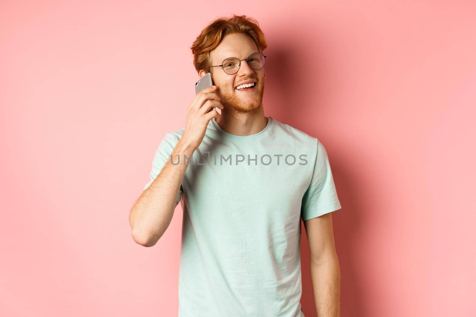 Handsomy hipster guy with red hair and beard talking on mobile phone, calling someone and looking happy, standing over pink background.