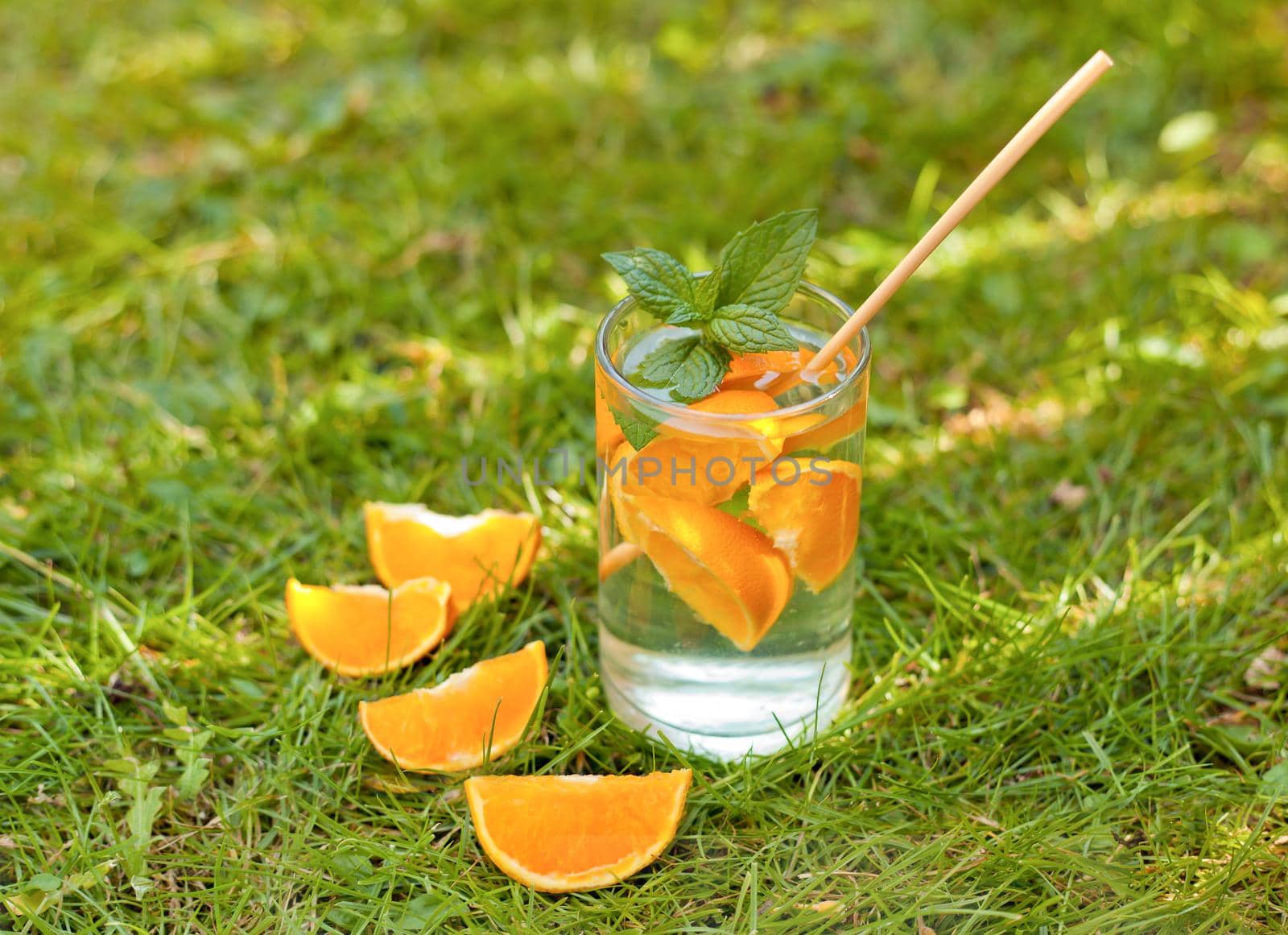 drinking glass with orange slices and water on the grass. cold lemonade drink with orange and mint in tall glass on green grass with eco straw