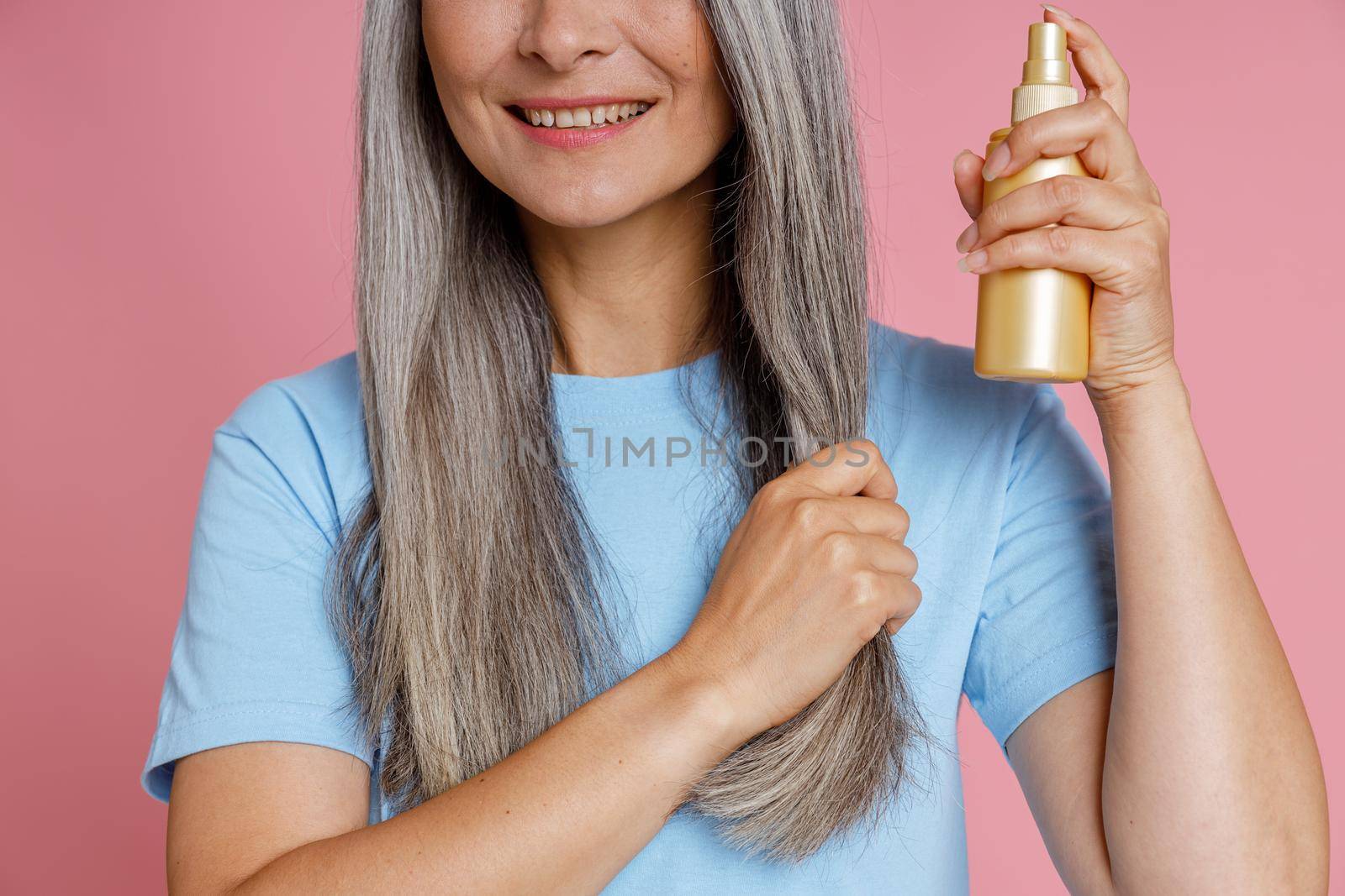 Smiling middle aged woman applies spray product onto grey hair lock on pink background by Yaroslav_astakhov