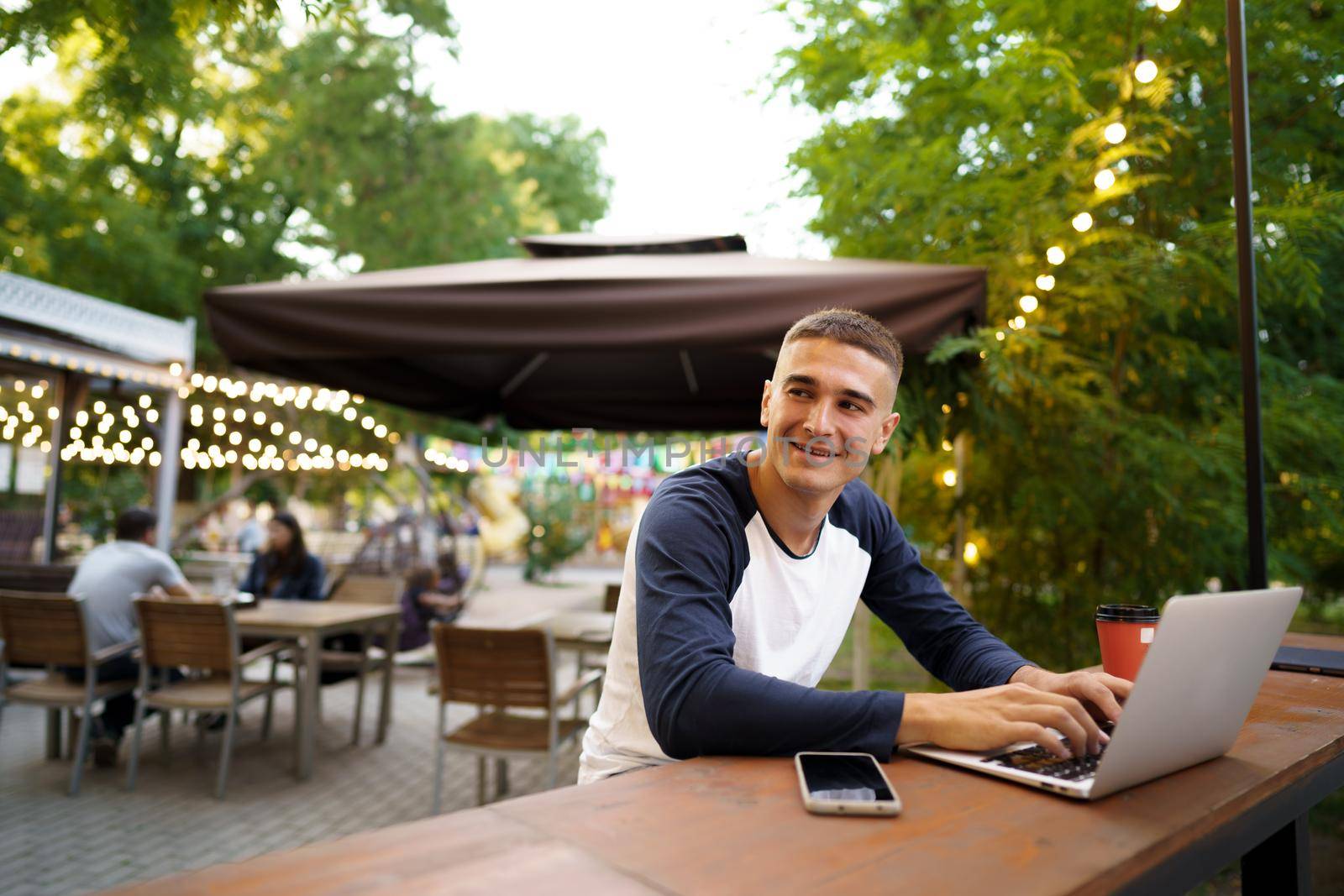 Young man sitting at table and typing on laptop keyboard while working in outdoor cafe by Fabrikasimf
