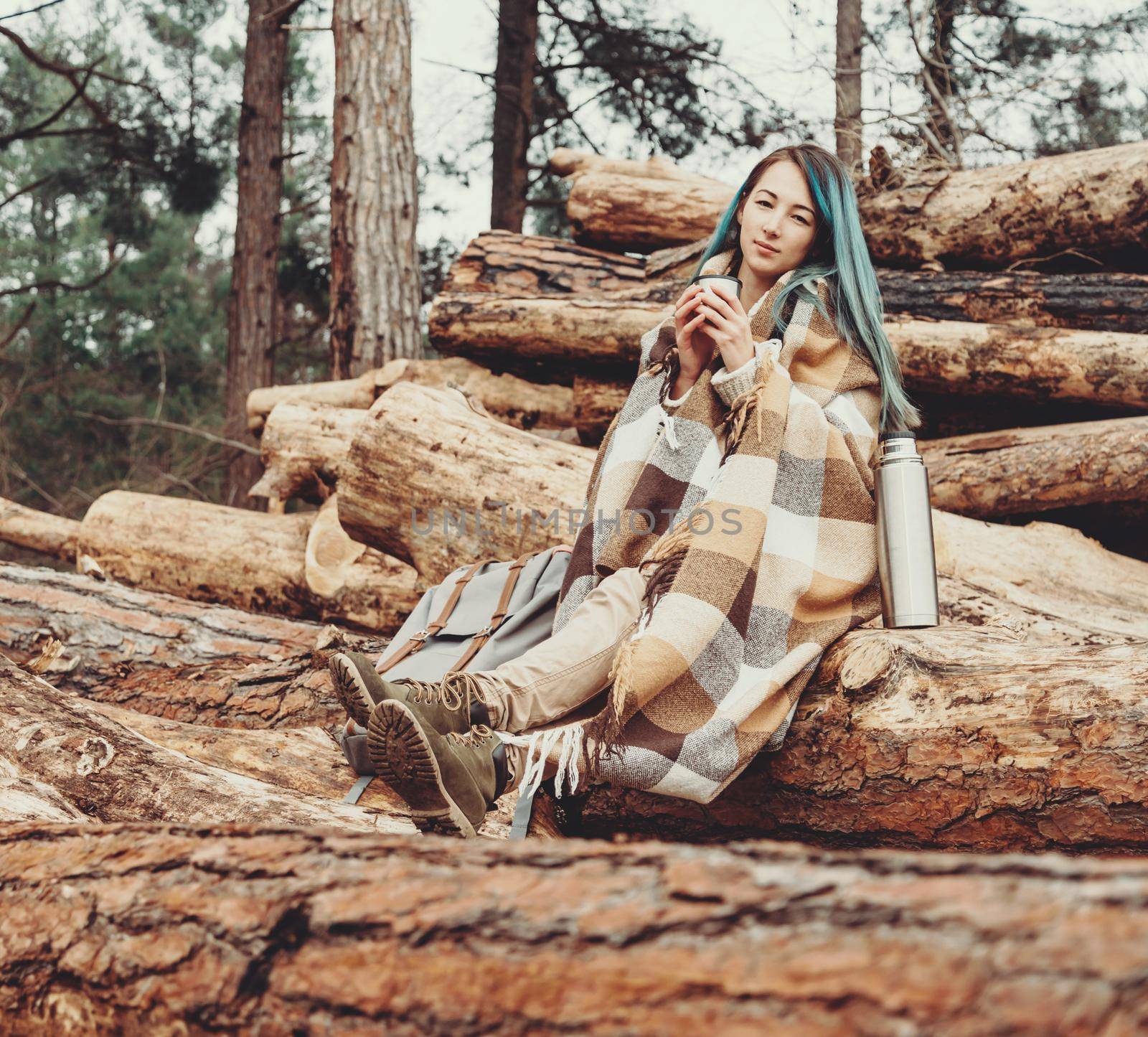 Young woman wrapped in plaid resting with cup of tea on stack of felled tree trunks in the woods