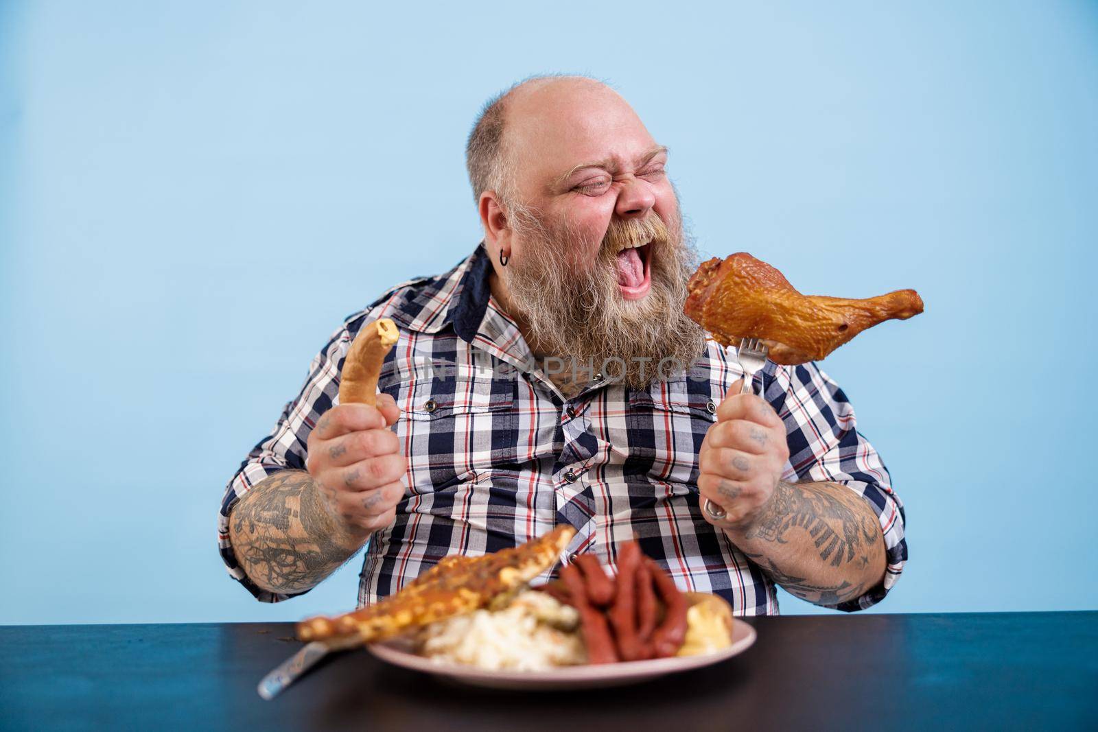 Hungry mature bearded man with overweight eats smoked chicken leg sitting at table with fat food on light blue background in studio