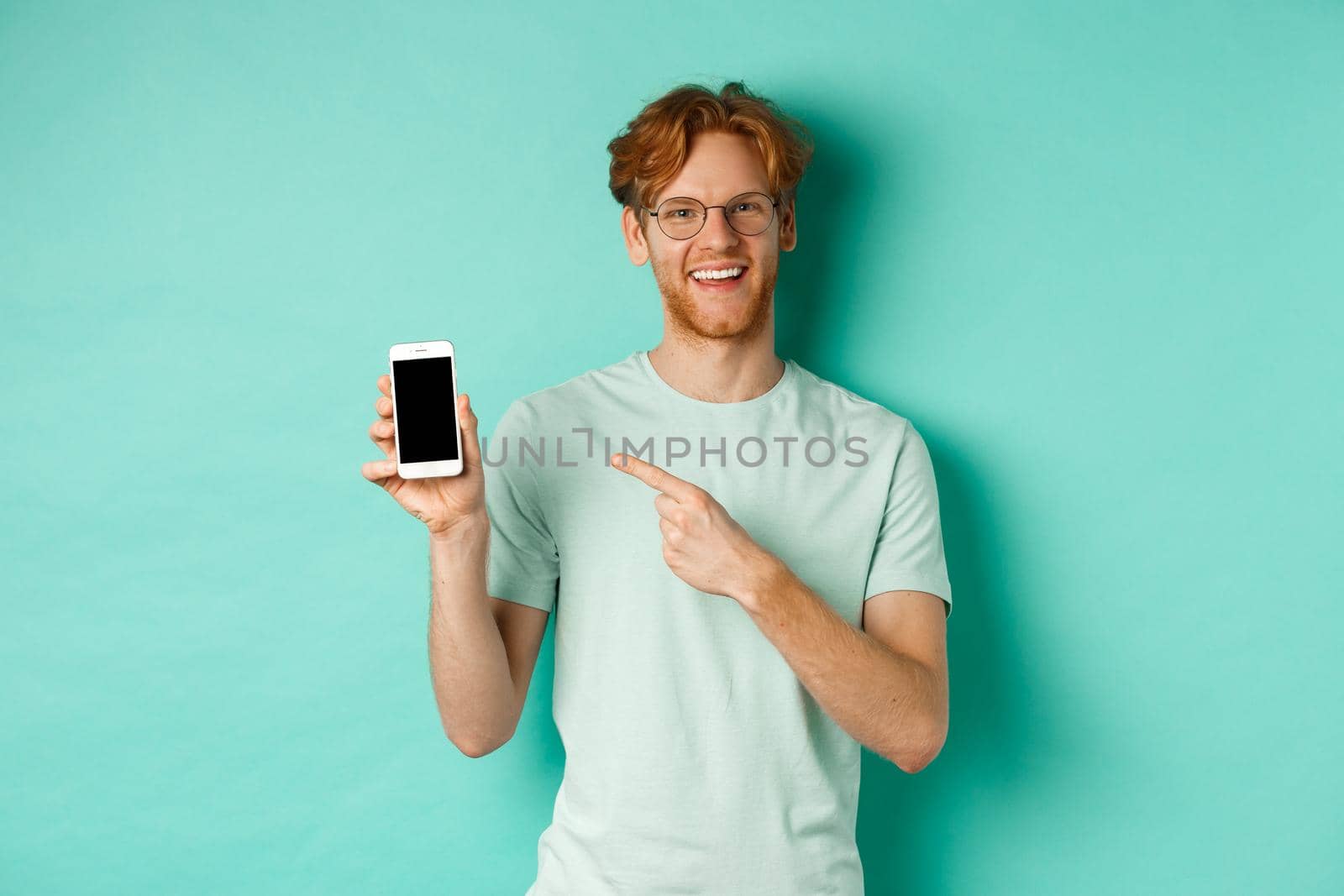 Attractive young man with red beard and hair pointing finger at blank smartphone screen, showing online promotion or app, smiling at camera, turquoise background by Benzoix