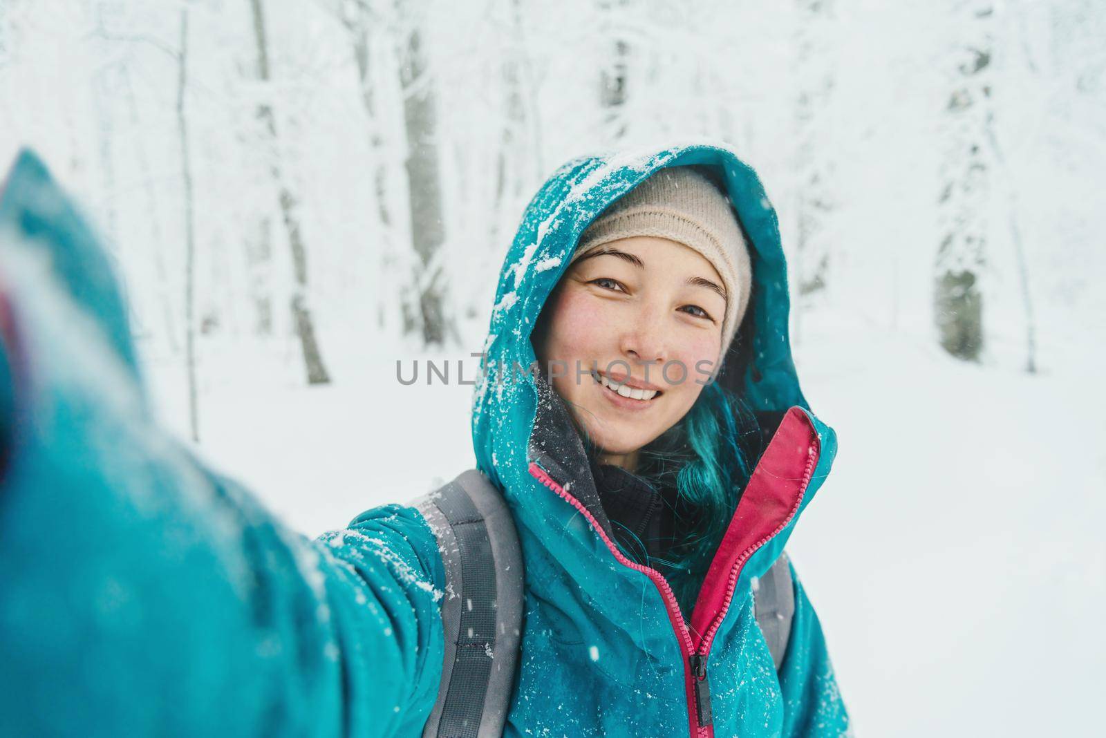 Smiling young woman doing selfie in snowy forest in winter, looking at camera