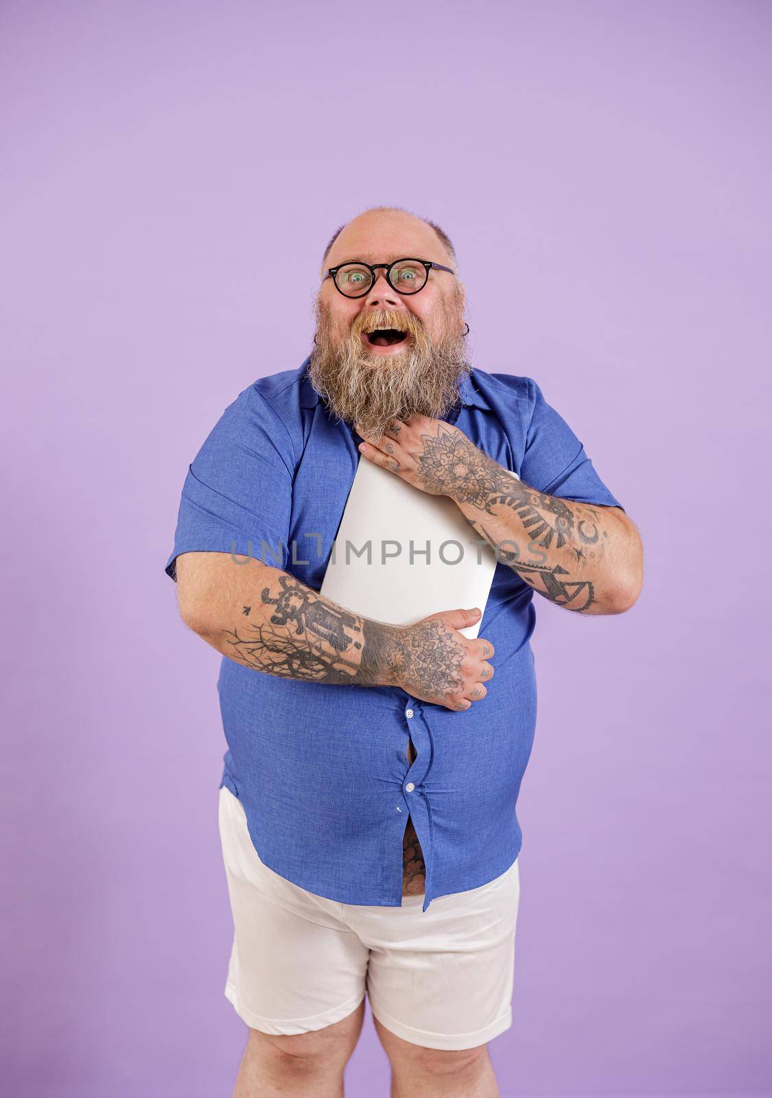 Excited bearded obese man in tight shirt embraces modern laptop on purple background by Yaroslav_astakhov