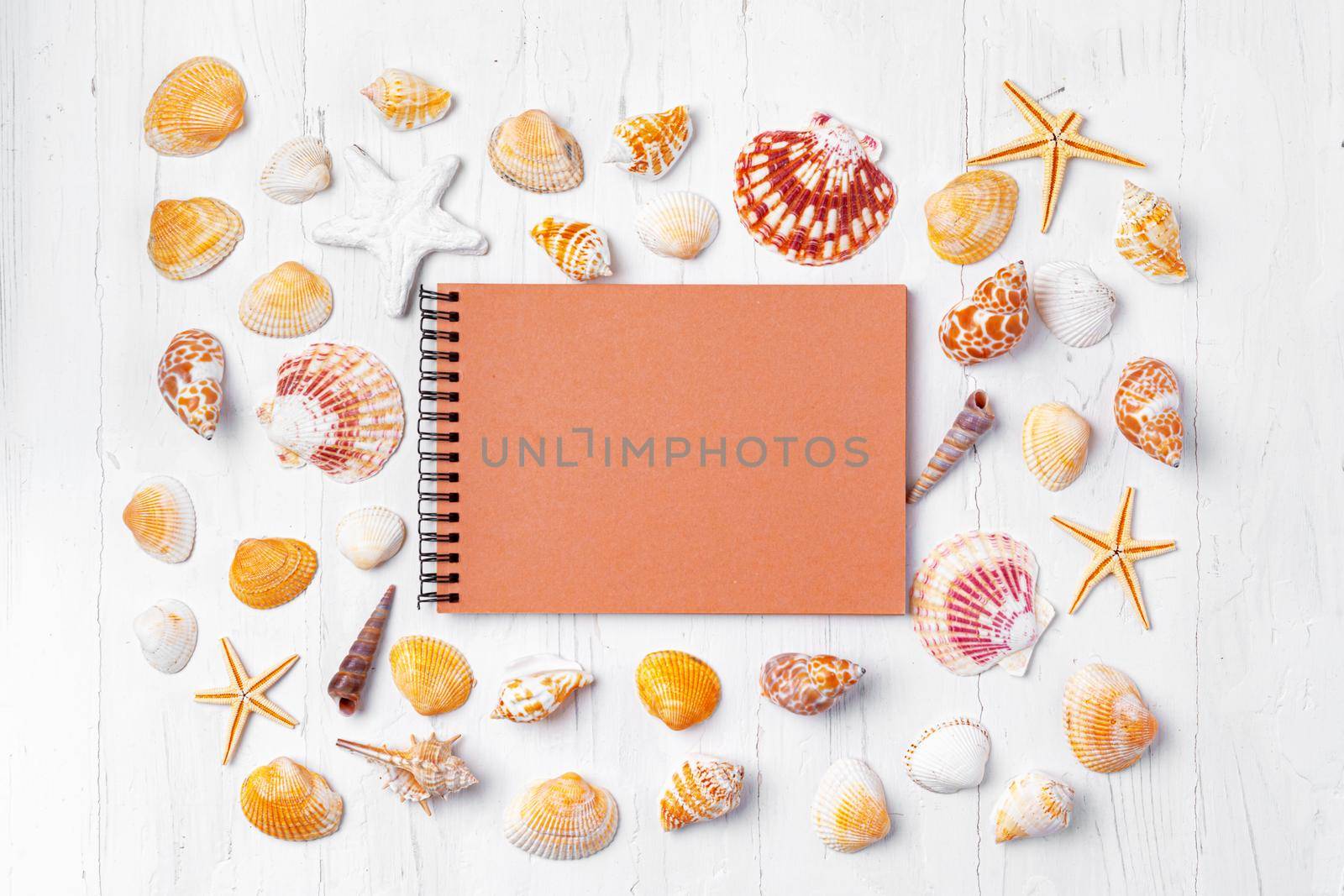 Open notepad with sea shells on white wooden background, top view