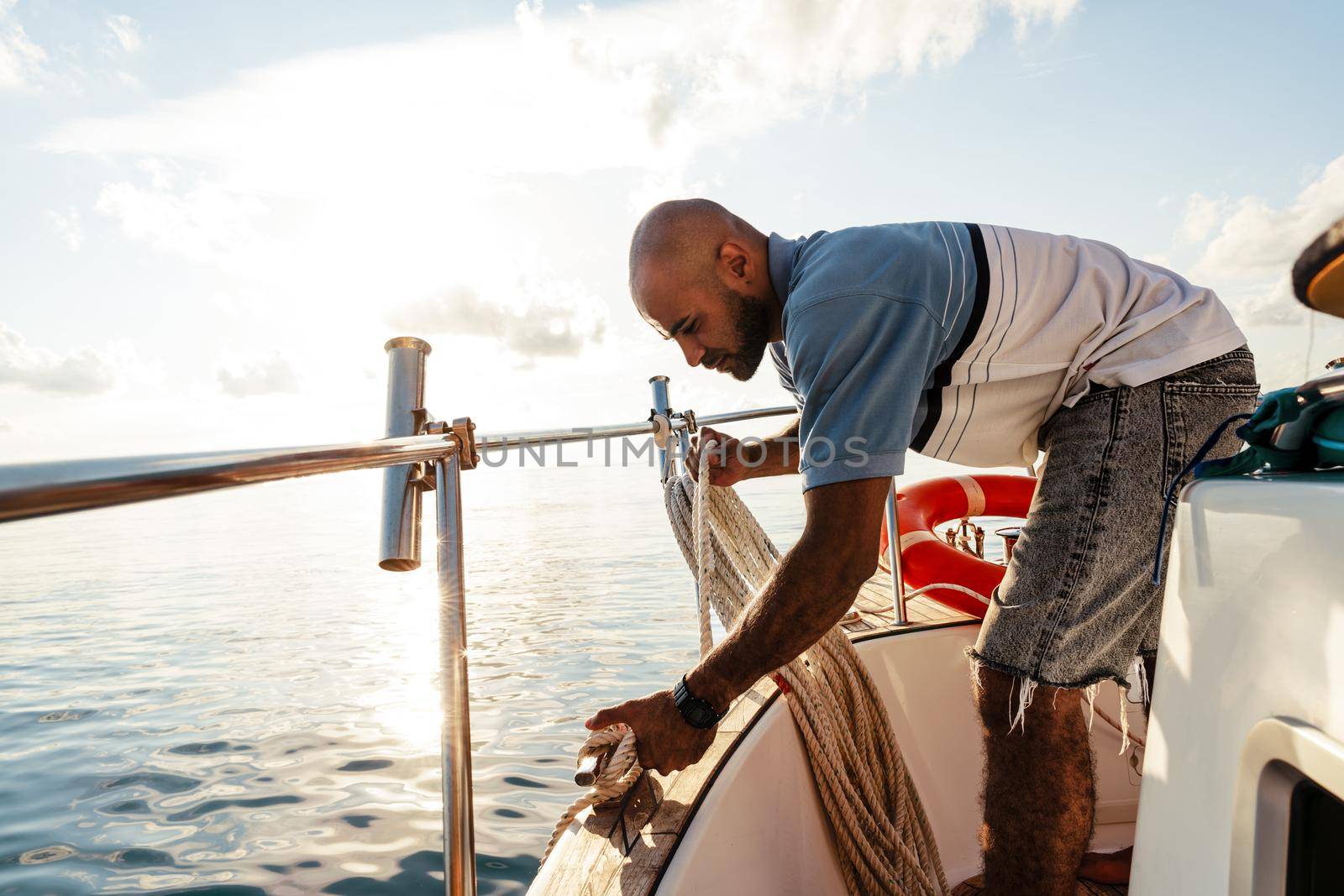 Young african american sailor tying ropes on sailboat in the sea on sunset, close up