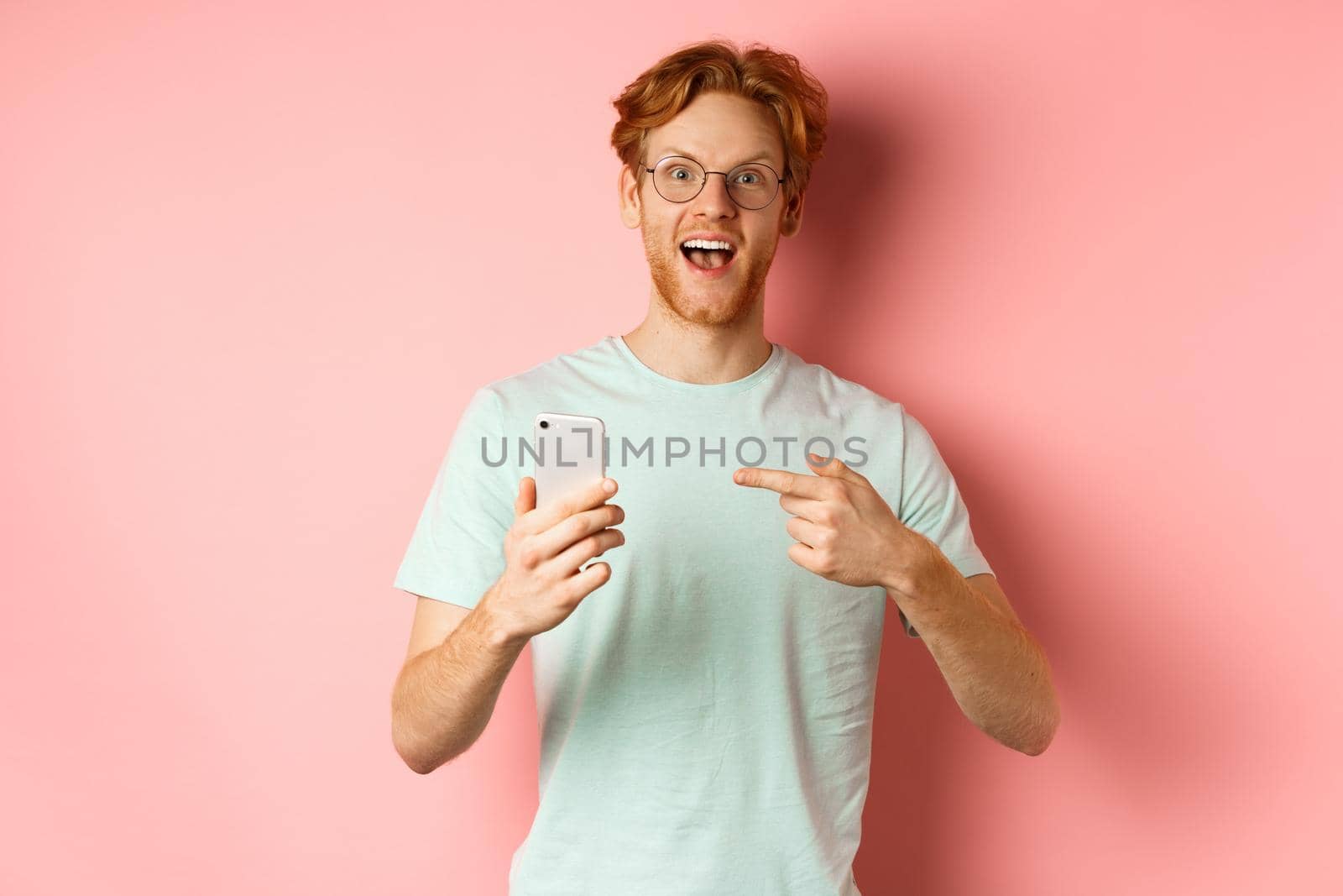 Cheerful guy talking about internet promo, smiling amazed and pointing finger at smartphone, standing over pink background.