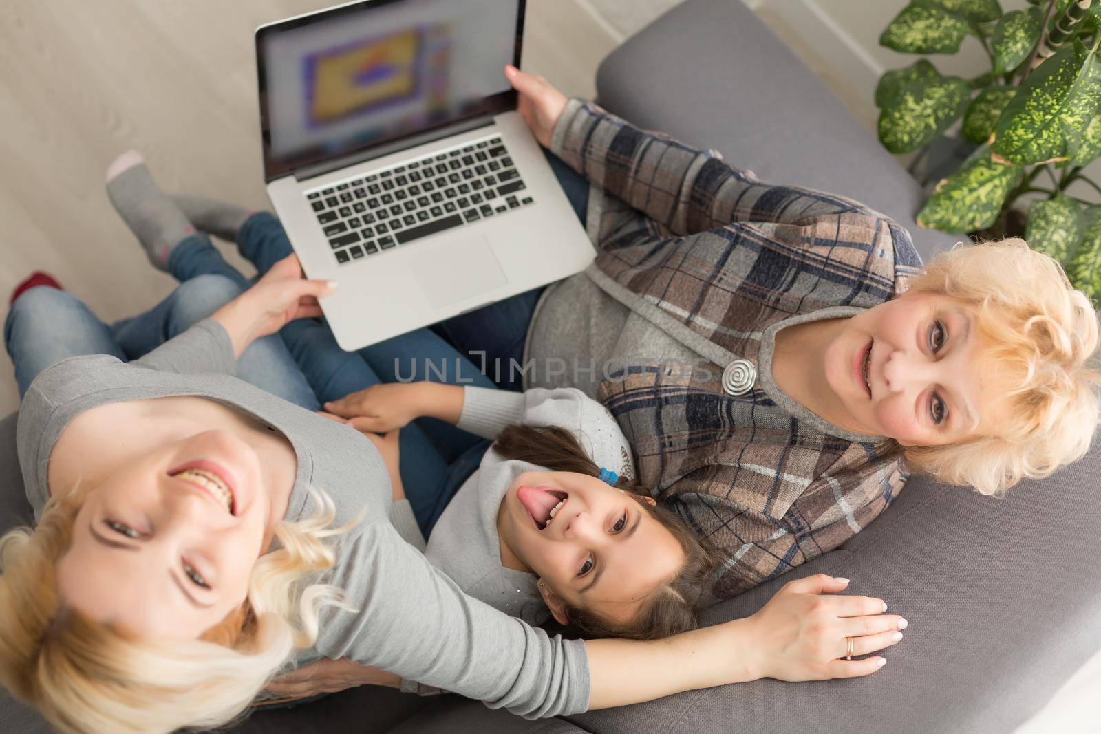 Happy three generations of women sit relax on couch laugh watching funny video on laptop, smiling positive females grandmother, mother and daughter have fun rest on sofa enjoy movie on computer