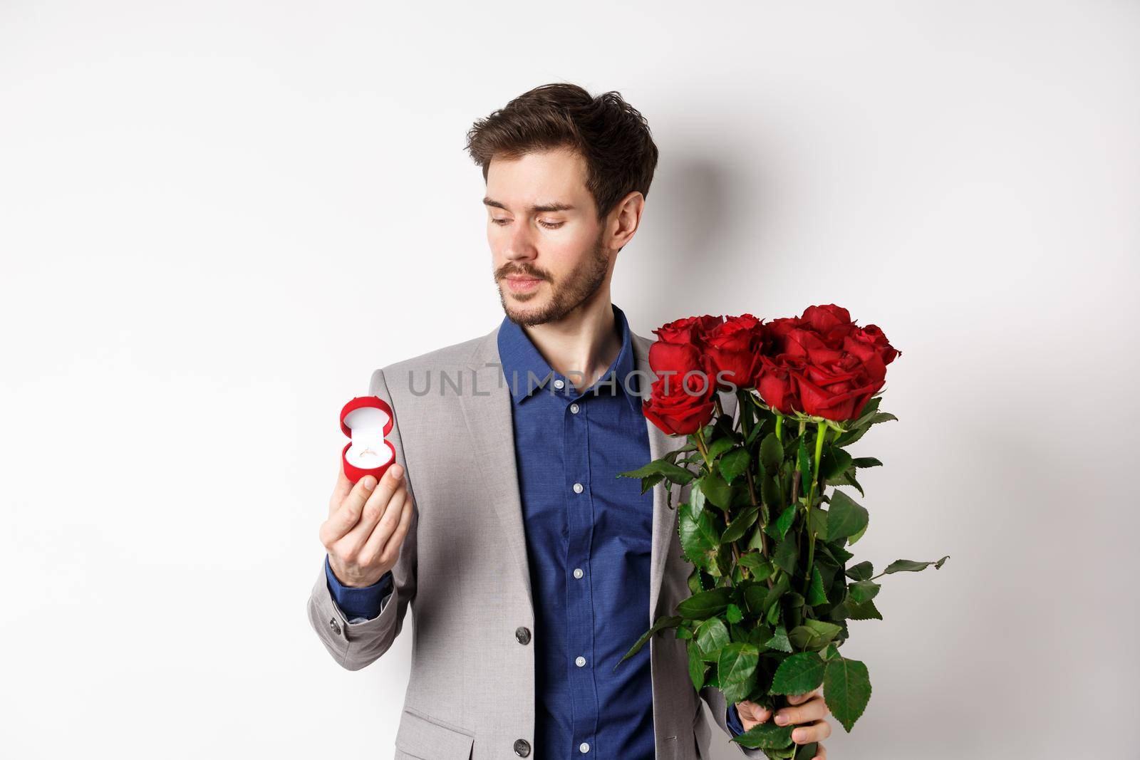 Handsome bearded man in suit looking at engagement ring, making surprise on lovers day, standing with red roses over white background.