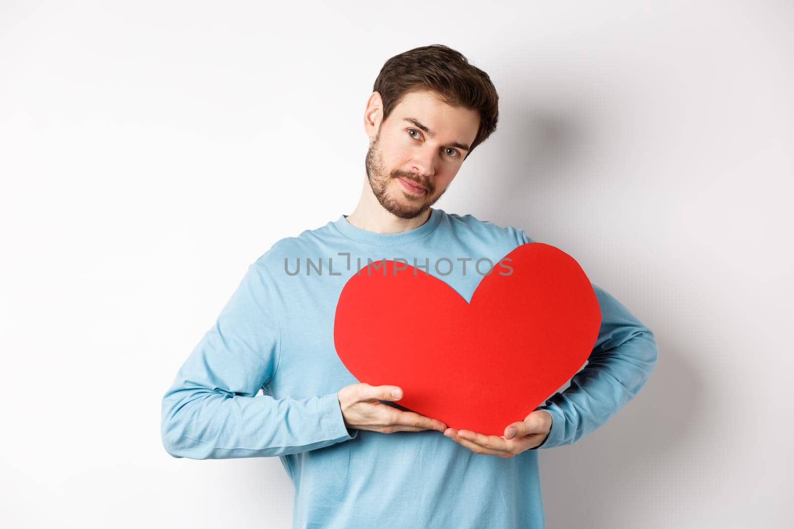 Romantic boyfriend making Valentines day surprise, holding big red heart cutout on chest and smiling with love, looking tender at camera, standing over white background.
