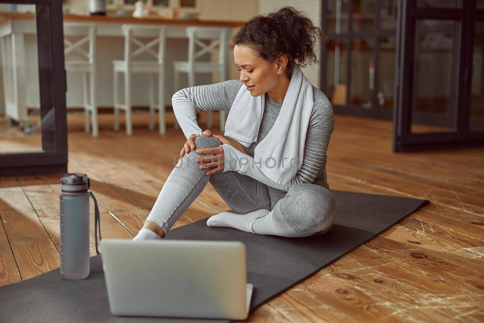 Female is sitting on mat and touching joint while suffering pain during video workout on laptop at home