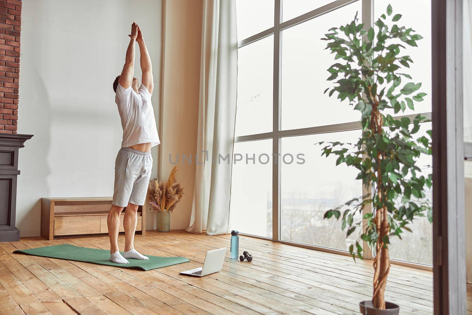 Male is practicing asana near window in living room while watching workout on notebook during quarantine