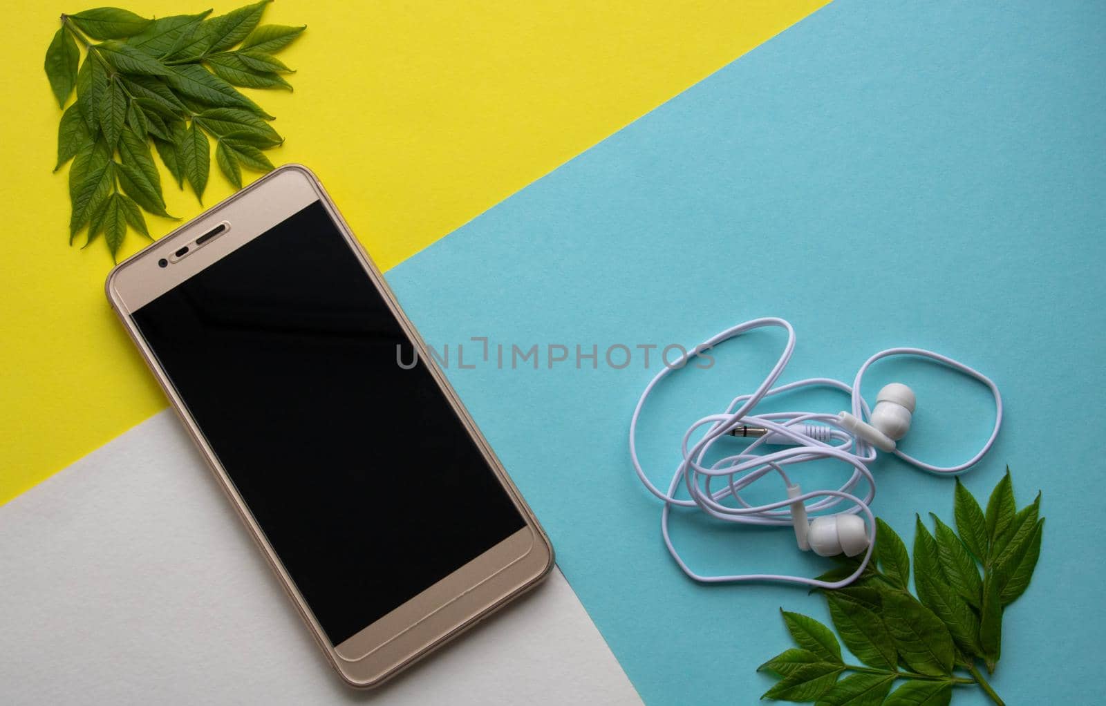 A Golden smartphone with a blank screen and headphones isolated on a white yellow and blue background with green leaves by lapushka62