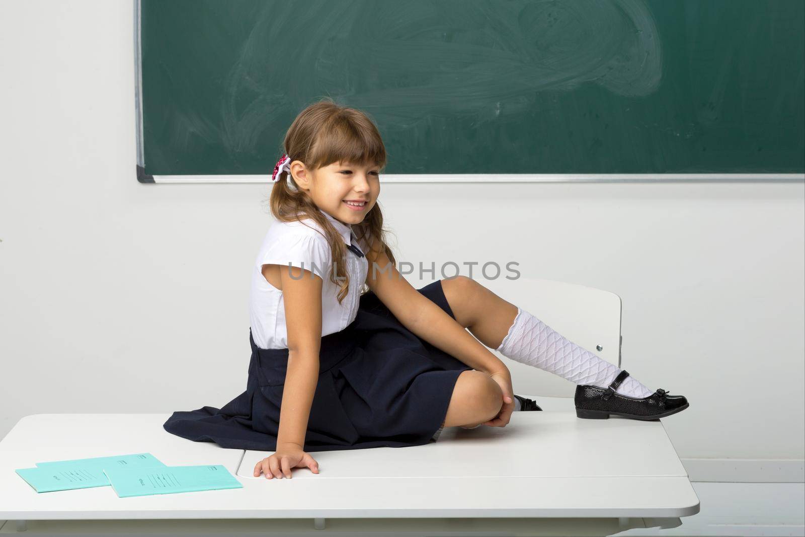 Happy school girl sitting on desk in classroom. Joyful girl student in white blouse and blue skirt relaxing after classes at school on background of blackboard. School and education concept
