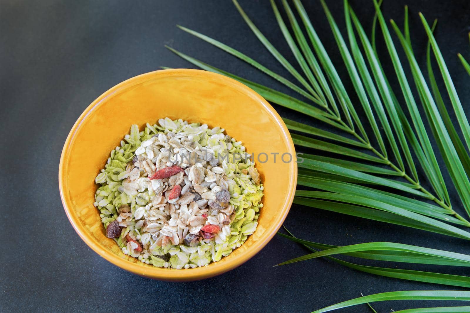 oatmeal with nuts and dried fruits in red bowl on black table near green palm leaf. Perfect whole grain breakfast as tasty organic food.