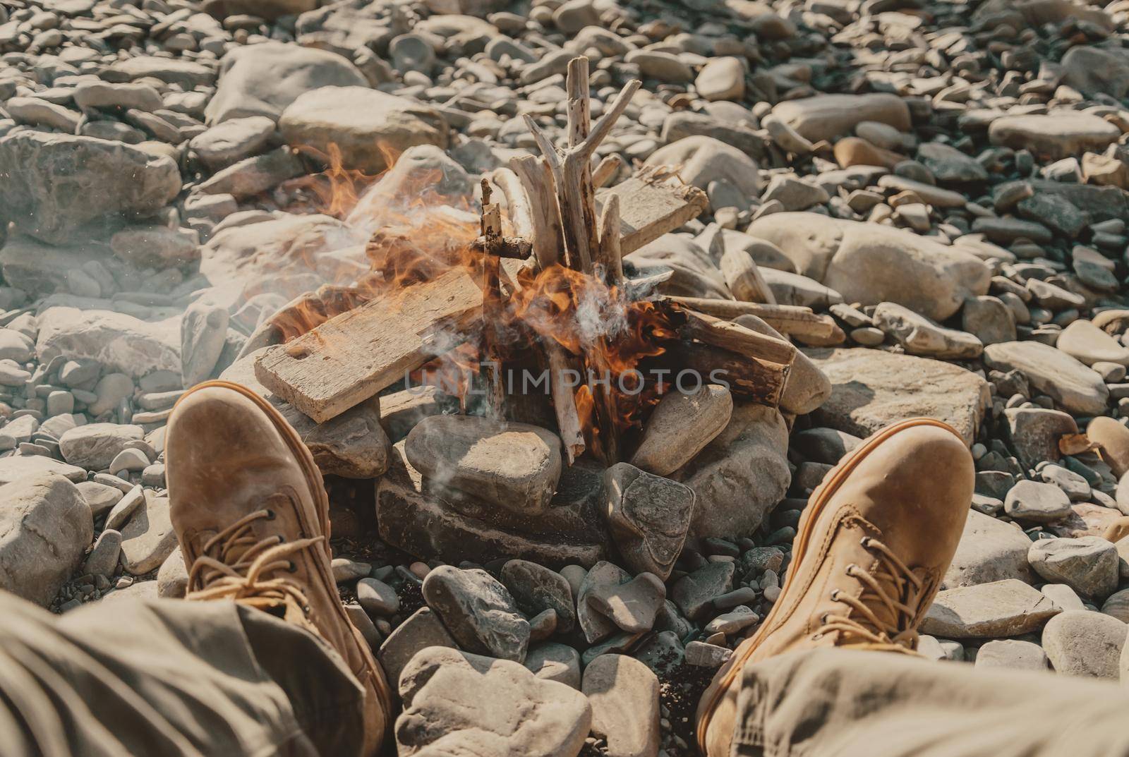 Young man sitting near the bonfire on pebble coast, view of legs, pov