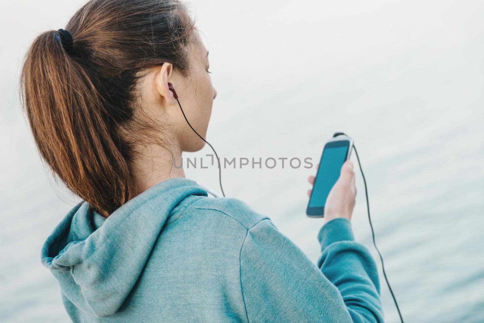 Fitness young woman in headphones listening music on her smartphone for workout on background of sea