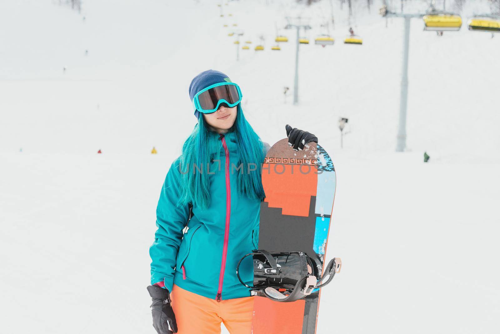 Young woman with blue hair wearing in protective sunglasses standing with snowboard on ski resort, looking at camera