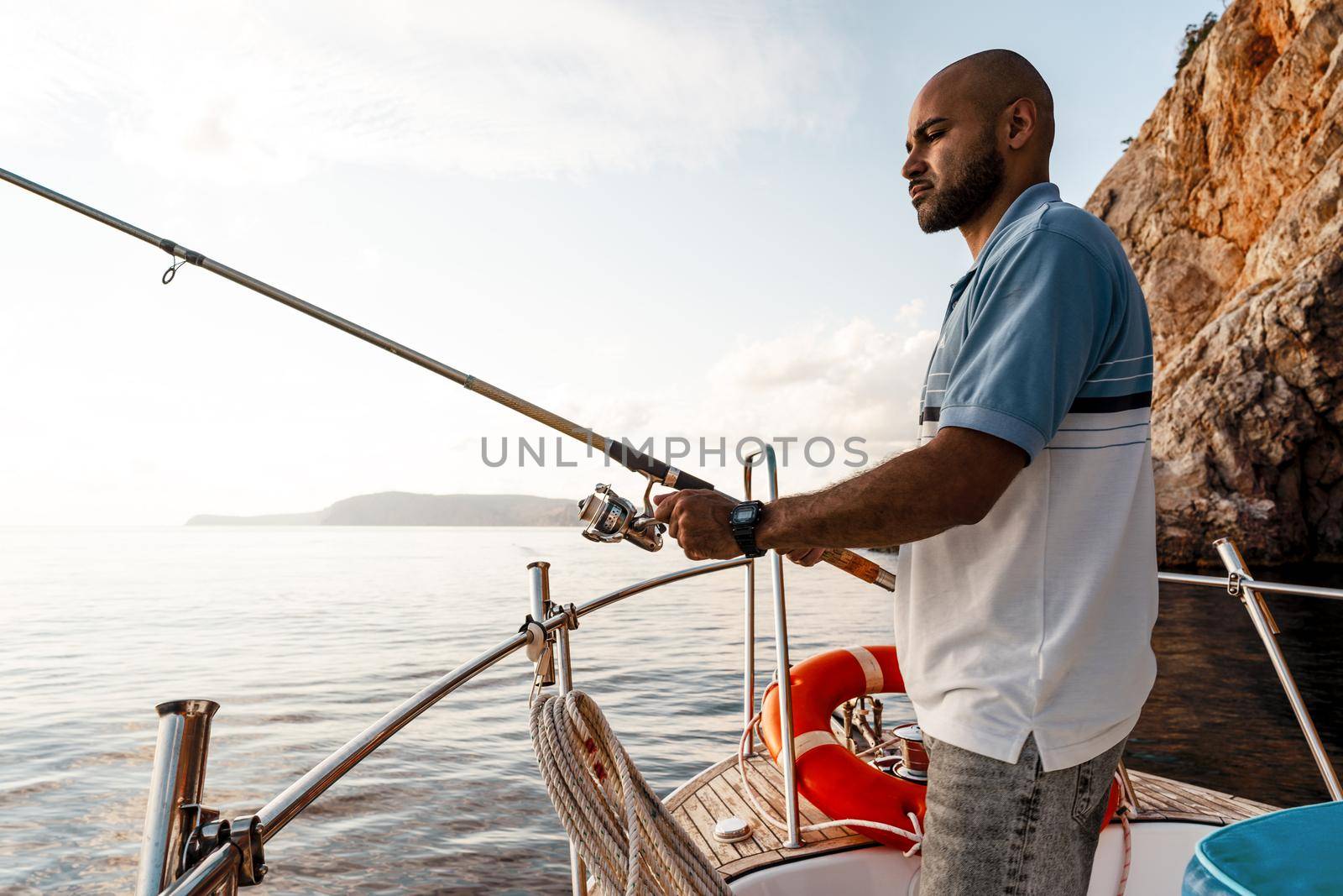 Young african american man standing with fishing rod on a sailboat fishing in open sea on sunset, close up