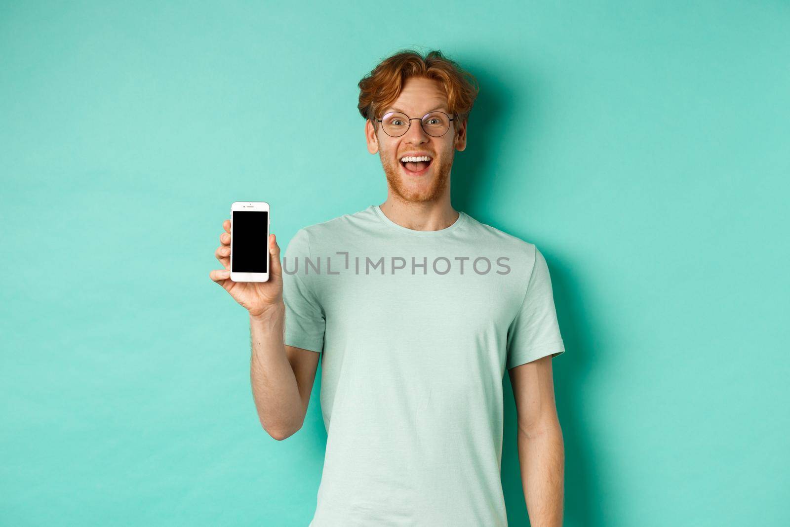 Online shopping. Cheerful redhead man in glasses and t-shirt showing smartphone screen and smiling, standing over mint background.